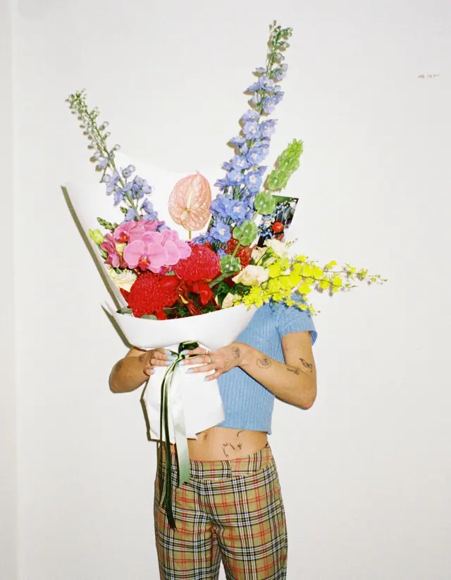 A wrapped bouquet of tall delphinium, red roses and yellow orchids held up against a white wall